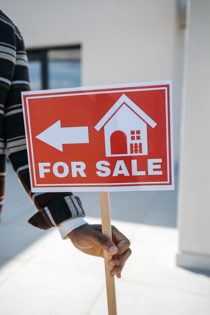 Close-up of a hand holding a For Sale real estate sign with a house icon outdoors.
