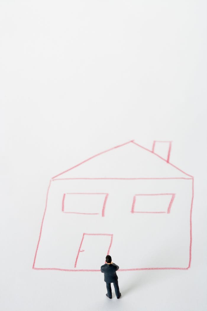 A miniature businessman stands before a simple house drawing on white background.